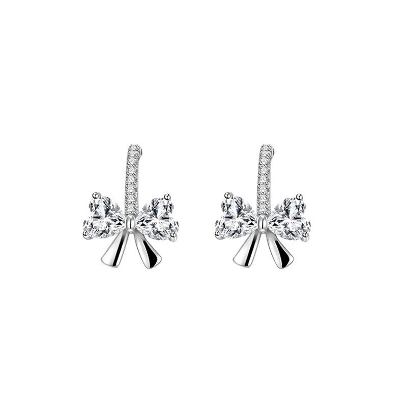 Sterling silver bow stud earrings with heart-shaped cubic zirconia stones, displayed on a soft beige background for an elegant gift look