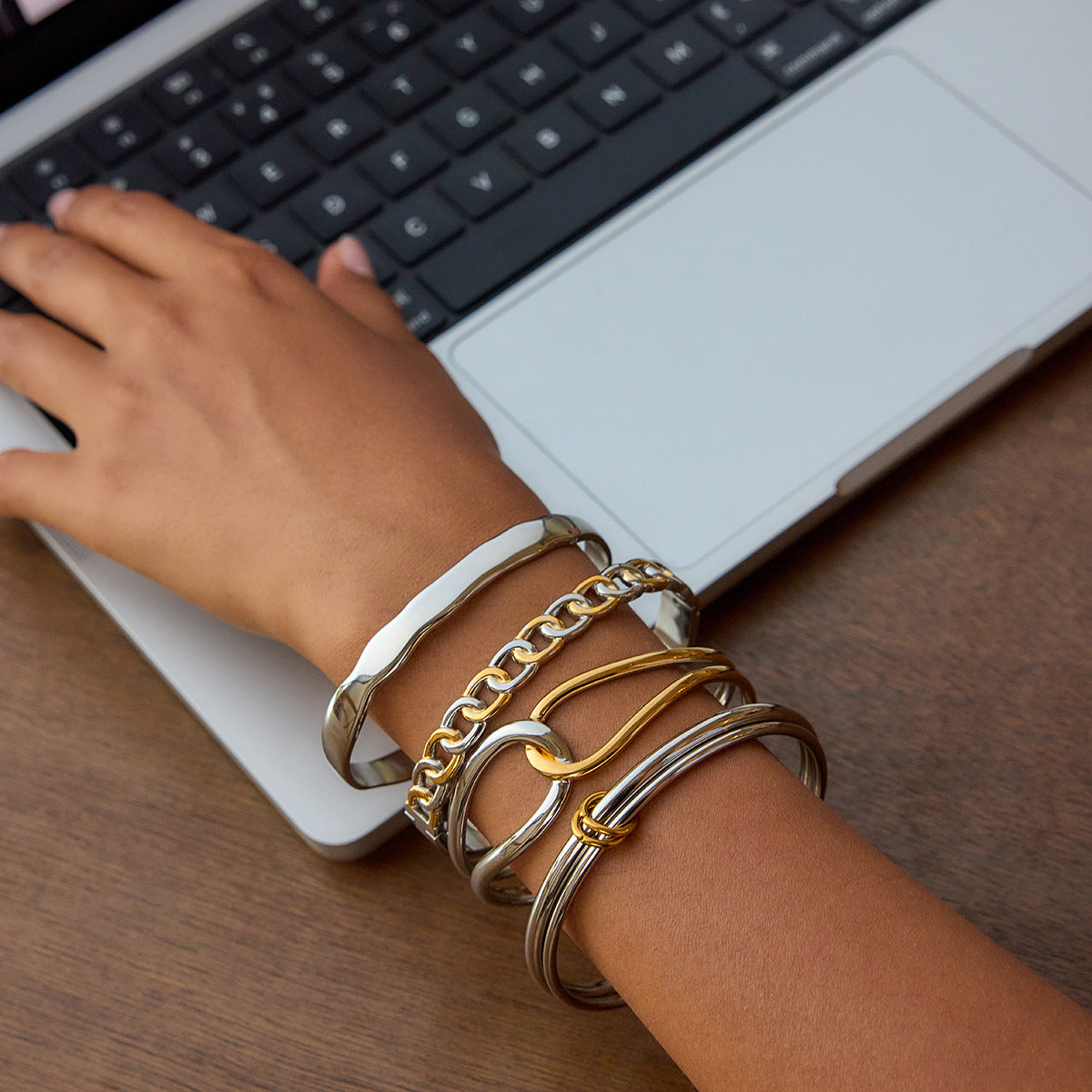 A stack of two tone gold and silver bangles styled with a dark feminine aesthetic, showcasing stainless steel bracelets worn on a wrist near a laptop — perfect for wholesale buyers or stylish women.