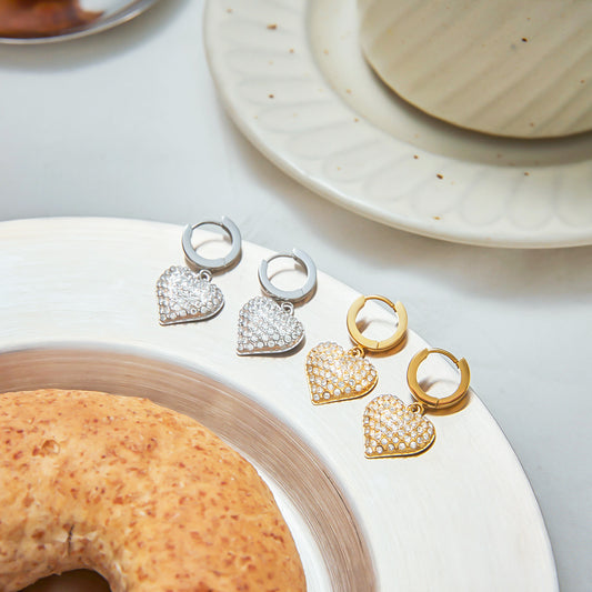 Back and front view of gold and silver heart zircon hoop earrings placed on a plate. Made of stainless steel with PVD gold plating, these drop earrings are durable and great for bulk jewelry wholesale listings.