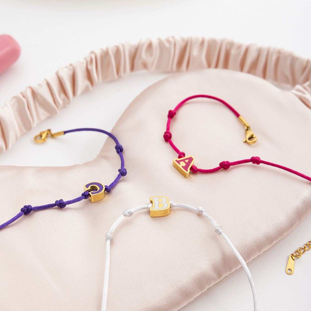 Wholesale enamel letter friendship bracelets with A B C initials in red, white, and purple string colors, displayed on soft satin pouch. Gold stainless steel adjustable bracelets perfect for personalized gifts for women.