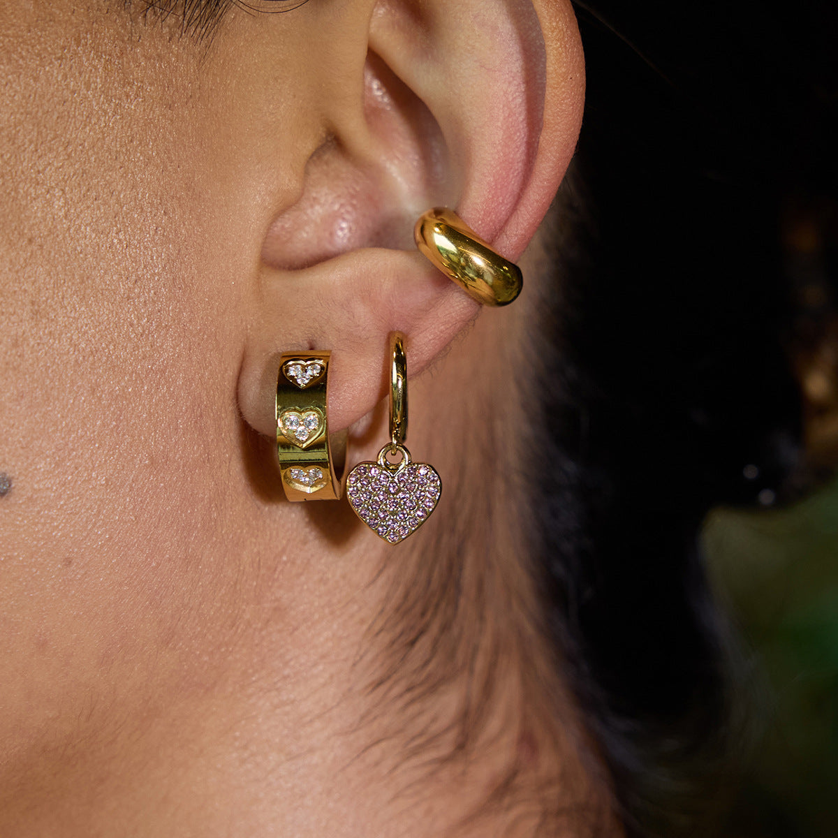 Model wearing stainless steel heart zircon earrings in gold, holding the silver version. These ladies fashion earrings feature PVD gold plating and are ideal for bulk orders or gifting collections.