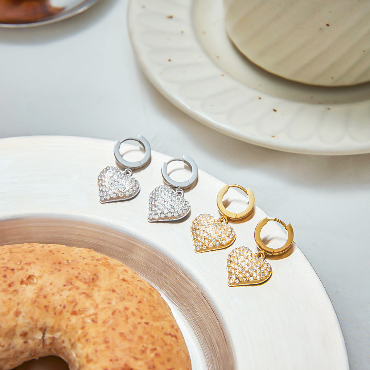 Back and front view of gold and silver heart zircon hoop earrings placed on a plate. Made of stainless steel with PVD gold plating, these drop earrings are durable and great for bulk jewelry wholesale listings.