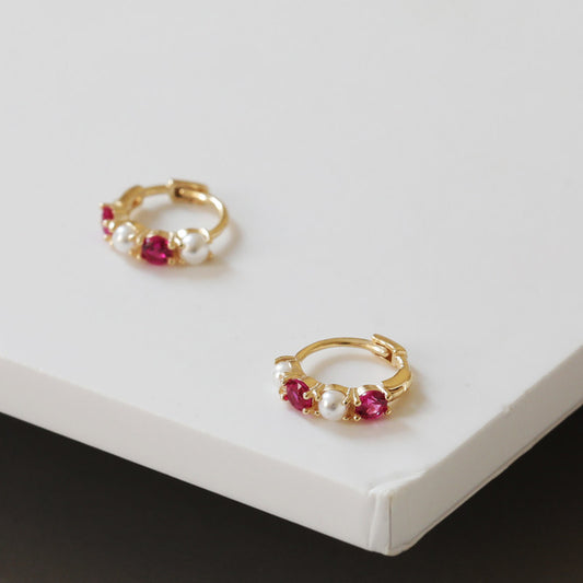 Close up of 18K gold plated sterling silver huggie earrings with ruby cubic zirconia and pearl details