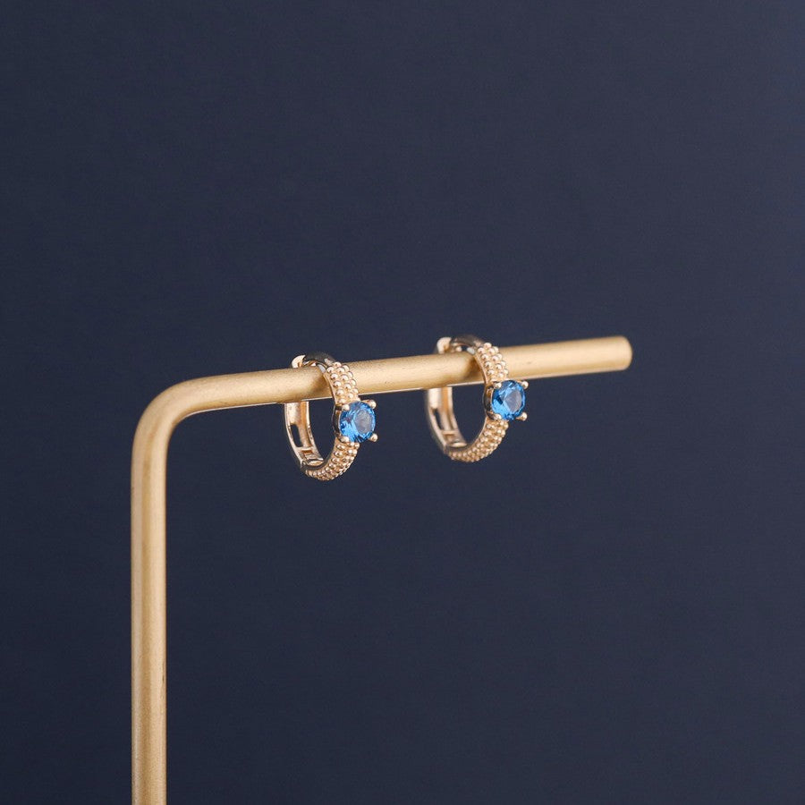 Blue sapphire cubic zirconia huggie hoop earrings displayed on jewelry stand with gold plated finish