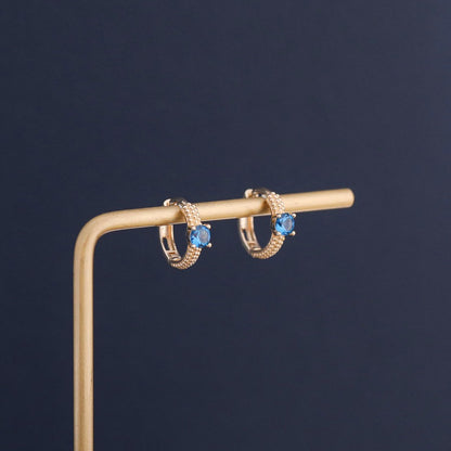 Blue sapphire cubic zirconia huggie hoop earrings displayed on jewelry stand with gold plated finish