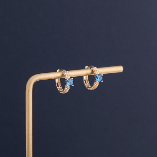 Blue sapphire cubic zirconia huggie hoop earrings displayed on jewelry stand with gold plated finish