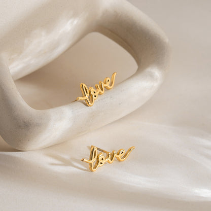 Gold stainless steel love letter stud earrings styled on shell display