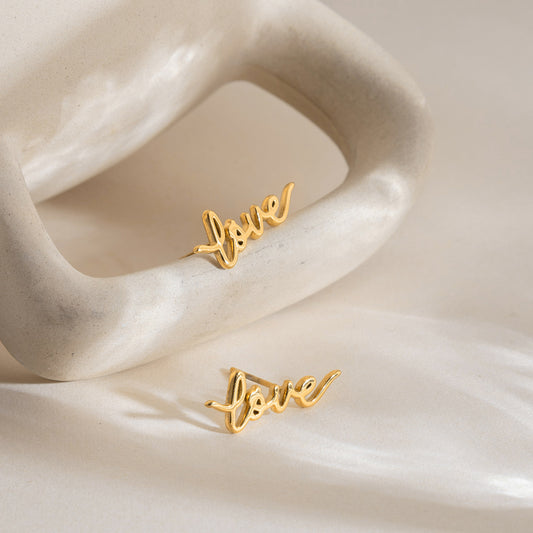 Gold stainless steel love letter stud earrings styled on shell display